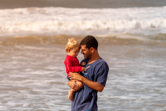 pai com filho no colo, com os rostos encostado, na praia.