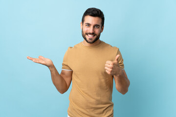 Fototapeta premium Young handsome man with beard over isolated background holding copyspace imaginary on the palm to insert an ad and with thumbs up