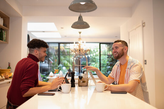 Happy gay male couple working from home at laptops on kitchen island
