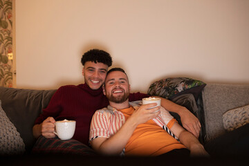 Happy affectionate gay male couple with hot cocoa watching TV on sofa