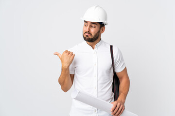 Young architect man with helmet and holding blueprints isolated on white background unhappy and pointing to the side