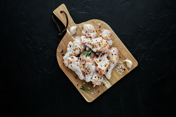 Raw chicken wings on cutting board with ingredients for cooking at black background. Fresh uncooked drumette or drumstick with garlic, chili pepper and rosemary. Raw meat. Top view with copy space.