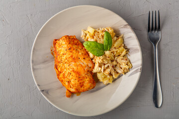 Baked chicken breast with salad on a plate next to a fork on a gray background.