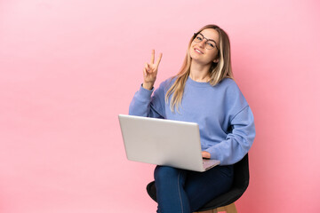 Young woman sitting on a chair with laptop over isolated pink background showing victory sign with both hands