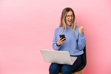 Young woman sitting on a chair with laptop over isolated pink background with phone in victory position