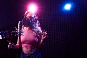 Beautiful blonde girl emotionally singing song in recording studio with professional microphone and headphones, creates new track album, vocal artist in pink blue neon light