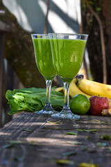 Dois copos com suco detox e ingredientes ao fundo: banana, couve, limão maça 