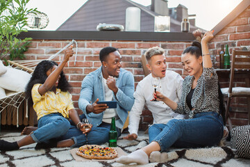 Four multiracial friends in stylish clothes having party on roof with alcohol, pizza and gadgets. Concept of happiness and real human emotions.