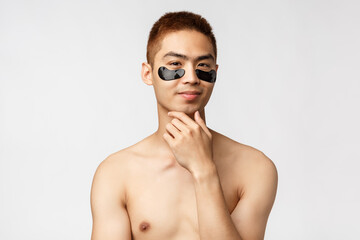 Fototapeta na wymiar Beauty, people and lifestyle concept. Thoughtful enthusiastic, handsome naked asian man wearing eye-patches and smiling, have an idea, thinking, consider apply skincare mask, white background
