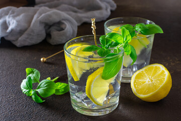 Two glasses of iced cocktail with lemons and basil on dark rustic background. Summer cold refreshing drink.