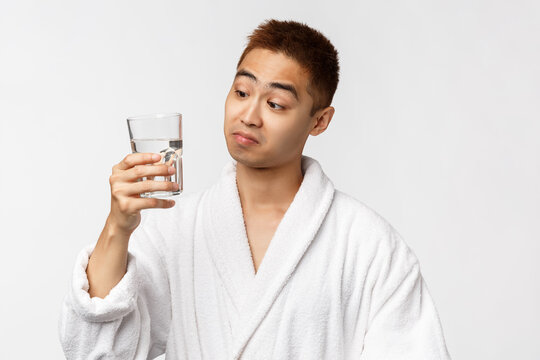 Beauty, Spa And Technology Concept. Not Bad Taste. Portrait Of Pleased Asian Man In Bathrobe Sipping Water From Glass And Look Satisfied Camera, Standing White Background