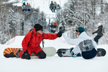 Couple of snowboarders gives a high five to each other while sitting on the snow tops of a slope...