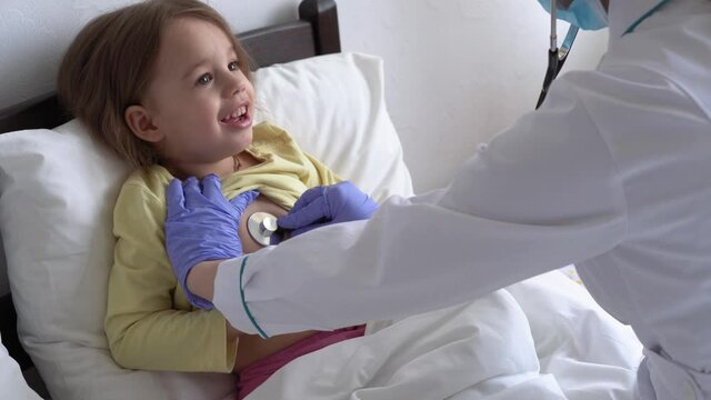 authentic cute caucasian cute young woman nurse or doctor pediatrician examine with statoscope minor preschool baby girl kid on white bed in hospital. medicine and health, pediatrics, covid-19 concept