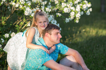 Father and daughter in the flowering white trees. A girl with blue eyes hugs the shoulders of a blond dad in a blue shirt. They sit before the branches of a flowering Jasmine.