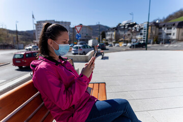 Fototapeta premium A young woman sits in a park bench alone and uses a smartphone. Virus pandemic times