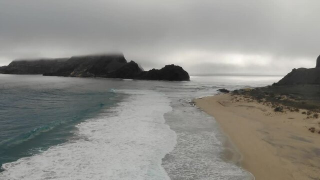 Aerial drone shot over beach with big rock shrouded in mist on background, Ponta da Calheta