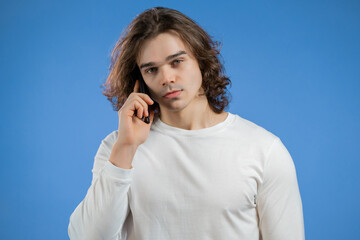 Fototapeta premium Young man speaks on phone. Guy holding and using smart phone. Blue studio background.