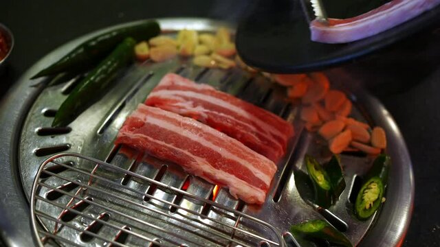 Chef hand cooking smoked grilled pork belly meat with garlic and green chili pepper on Korean iron BBQ grill pan in restaurant. Traditional Korea style food barbecue pork belly steak on charcoal grill