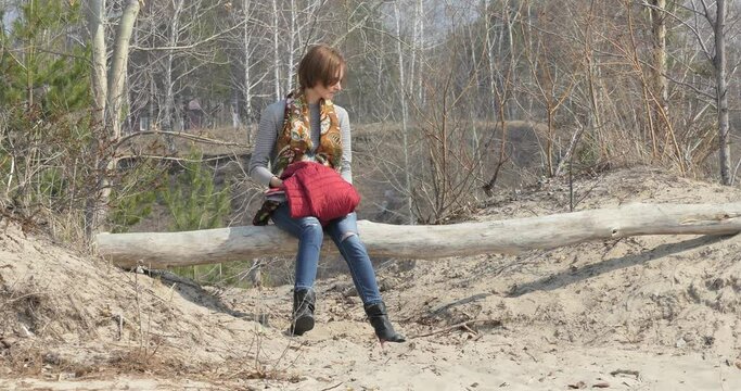 A woman in self-isolation in jeans sits on a log on a sandy beach against the background of spring trees that are not yet covered with leaves and looks around.