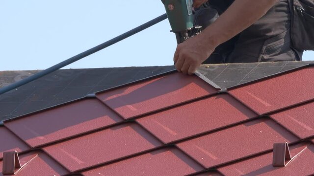 CLOSE UP: Unrecognizable roofer lays aluminium tiles with the help of a nail gun. Construction site builder helps lay the roofing atop a modern house. Contractor uses power tools during roofing works.