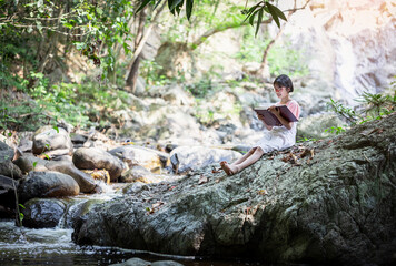 Asian little girl with glasses sitting on stone reading a books near forest waterfall