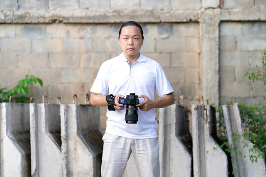 Asian man serious looks at the camera while holds th mirrorless camera medium format in the hand on the construction concret wall background.