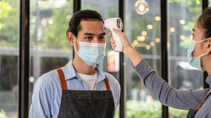Young Asian restaurant or cafe waiter service staff wearing face mask had body temperature screen check by colleague before enter workplace for prevent coronavirus infection to customer.
