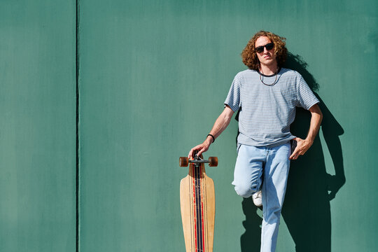 a young man with curly long hair and sunglasses with a longboard or skate with a green wall behaind him