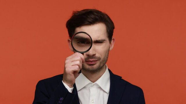 Portrait of bearded businessman sees through magnifying glass, trying to find something small, big man eye, funny male detective using loupe. Indoor studio shot isolated on orange background