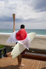 Man standing holding a flaming torch used in a large sporting event, with a surfboard and a Japanese Flag looking at the Pacific Ocean in Chiba Japan where a big surfing competition is held.