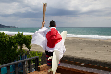 Man standing holding a flaming torch used in a large sporting event, with a surfboard and a Japanese Flag looking at the Pacific Ocean in Chiba Japan where a big surfing competition is held.