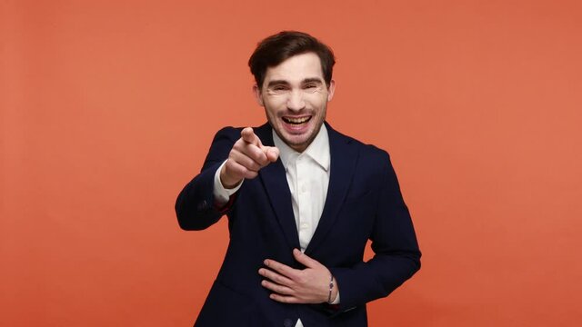Bearded businessman bursting into laughing and pointing to camera with finger, taunting somebody with loud laughter, making fun towards you. Indoor studio shot isolated on orange background.