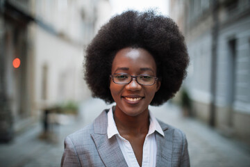 Portrait of smiling african woman in eyeglasses and business suit smiling and looking at camera while standing on city street. Concept of success, confidence and business people.