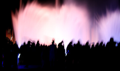 Naklejka na ściany i meble Silhouettes of people in motion near a colored fountain at night.