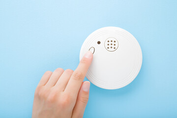 Fototapeta na wymiar Young adult woman finger pushing button of new white plastic smoke alarm. Light blue table background. Pastel color. Closeup. Safety concept. Front view.