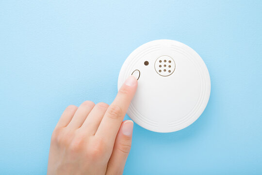 Young adult woman finger pushing button of new white plastic smoke alarm. Light blue table background. Pastel color. Closeup. Safety concept. Front view.