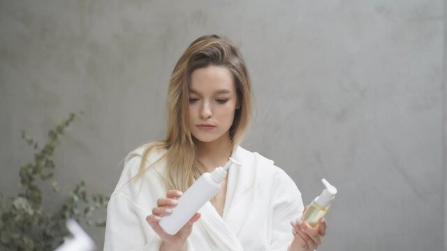 Thoughtful female selects, compares cosmetics, which one will be better, stand in bathrobe in bathroom at home, in front of mirror. at home.