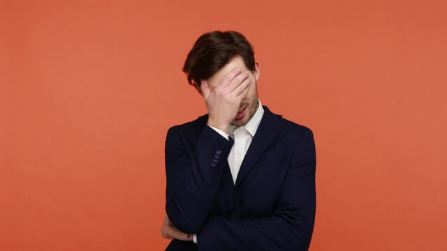 Unhappy bearded man slapping hand on forehead, doing facepalm gesture, looking at camera repentance, regret for mistake failure. Indoor studio shot isolated on orange background