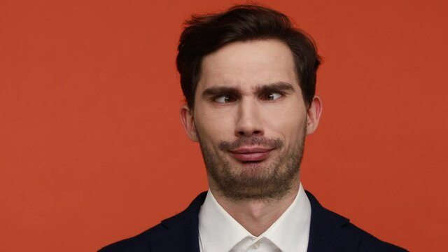 Crazy childish young businessman in official suit looking around with crossed eyes, making silly brainless face, looks stupid, aping as fool. Indoor studio shot isolated on orange background