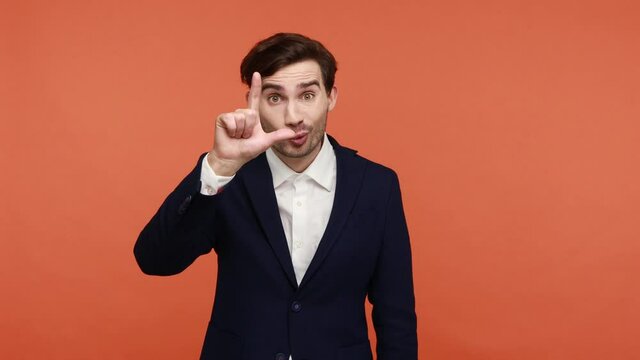 Bearded young guy standing with L finger sign and pointing at camera, saying you are loser, blaming for defeat, teasing mocking for failure. Indoor studio shot isolated on orange background.