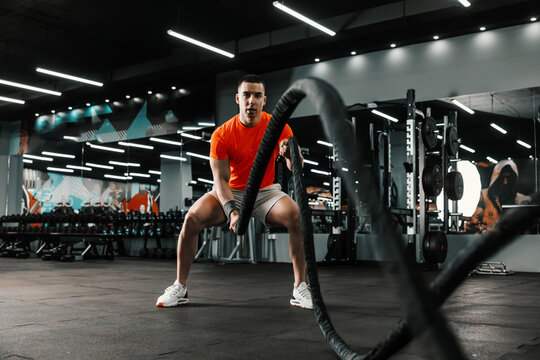 An energetic sportsman is doing heavy cardio cross-fit training with battle ropes in an indoor gym with a black background and a big mirror. Functional training, sports lifestyle