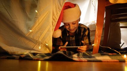 Happy little boy in Santa hat lying in DIY kid's tent celebrating Christmas using smartphone, having video call. Christmas fun at home.