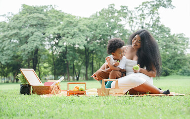 African family mother and little son sitting on mat picnic together at green park. Mom is looking at her child with love while he enjoy eating apple