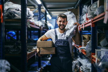 Smiling bearded tattooed hardworking blue collar worker in overalls holding boxes and bag and relocating them while walking in storage of import and export firm.