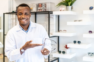 Eyesight And Vision Concept. Portrait of smiling african american standing at optics store and pointing at showcase and rack with many eyewear and specs frames. Guy offering spectacles