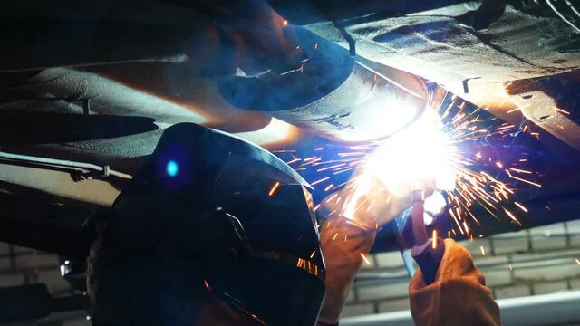 Car service worker welds and repairs an exhaust pipe of a car. Close-up welding work on a lifted car. View from the back. Mechanic welds the bottom of the car.