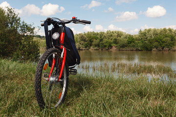 The backpack is hung on the handlebars of a bicycle. Red bicycle on a background of lake and bushes