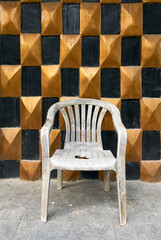 Close-up of an old dirty and broken white plastic chair standing on a floor in front of a gold and black checkered wall of closed-down establishment