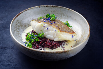 Modern Style traditionell gebratenes Skrei Kabeljau Fisch Filet mit Baby Brokkoli und schwarzen Reis in Zitronen Creme Sauce serviert als close-up in einer Nordic Design Schale