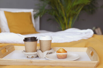 Two coffee cups and a cupcake on a white bed with an orange bedspread in a hotel bedroom banner. Romantic weekend for a couple with breakfast in bed on Valentine's Day. 
